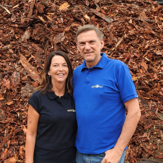 Ein Mann und eine Frau stehen gemeinsam vor einem großen Haufen Holzspäne, beide tragen passende schwarze und blaue Shirts.