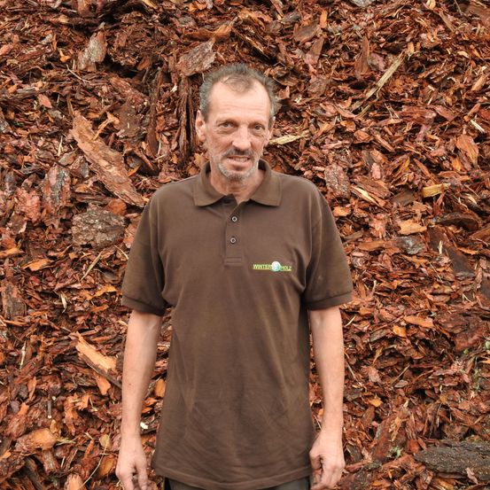 Ein Mann in einem braunen Polohemd steht vor einem großen Haufen brauner Holzspäne und lächelt.