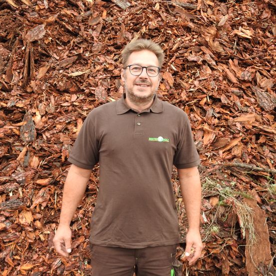 Ein Mann mit Brille und braunem Hemd steht vor einem großen Haufen brauner Holzspäne.