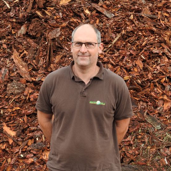 Ein Mann mit Brille und einem braunen Poloshirt mit dem Wort Holz steht vor einem großen Haufen Holzspäne.
