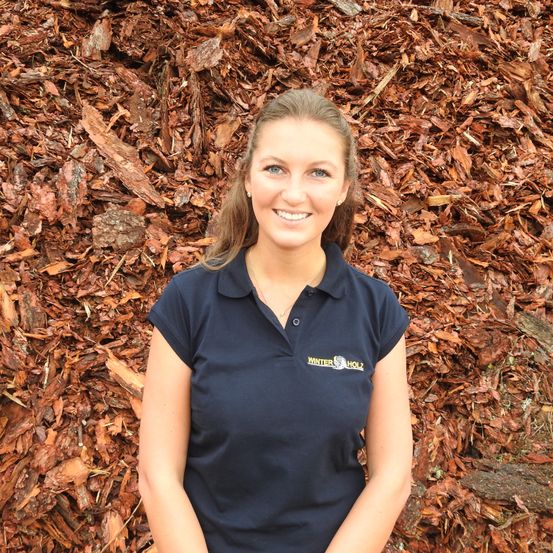 Eine Frau steht vor einer Wand aus braunen Holzspänen und lächelt. Sie trägt ein dunkelblaues Shirt mit einem Logo auf der linken Brust.