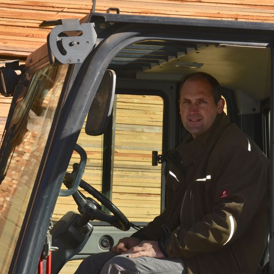 Ein Mann in einer braunen Jacke sitzt in einem Fahrzeug mit einem Lenkrad und einem Armaturenbrett und lächelt. Das Fahrzeug ist neben einer Holzstruktur geparkt.
