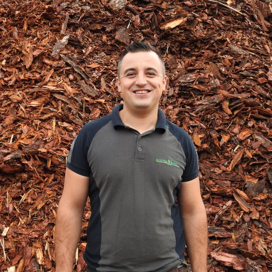 Ein Mann in einem grauen und blauen Poloshirt steht vor einem großen Haufen Mulch. Er lächelt und scheint für ein Foto zu posieren.