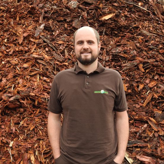 Ein Mann in einem braunen Polohemd steht vor einem großen Haufen von Holzspänen und lächelt.