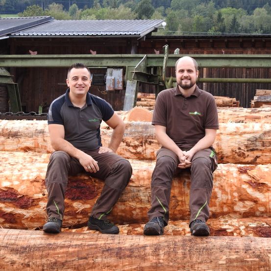 Zwei Männer sitzen lächelnd auf einem Haufen Baumstämme. Hinter ihnen steht ein Holzgebäude mit einem grünen Metalldach. Bäume und Berge sind im Hintergrund.