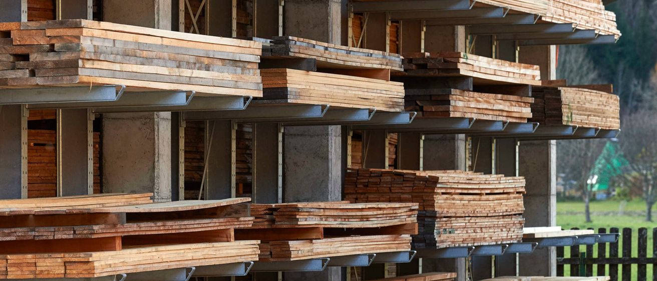 Ein Lagerraum mit Holzbrettern, die auf Metallregalen gestapelt sind. Die Bretter sind ordentlich angeordnet und zeigen verschiedene Größen und Dicken. Die Umgebung wirkt industriell.