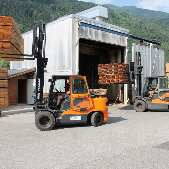 Zwei Gabelstapler stehen vor einem Lagerhaus mit Holzstapeln im Inneren. Die Gabelstapler sind orange und eine Person fuhrt jeden von ihnen. Im Hintergrund sind Berge zu sehen.