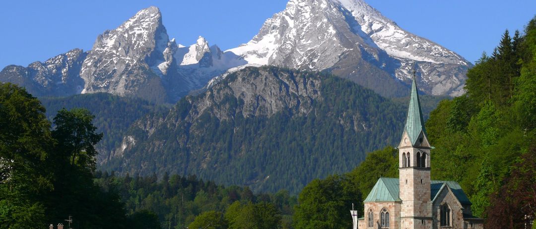 Ein malerisches Dorf in den Bayerischen Alpen, mit einer Kirche und Häusern unterhalb schneebedeckter Berge unter einem klaren blauen Himmel.
