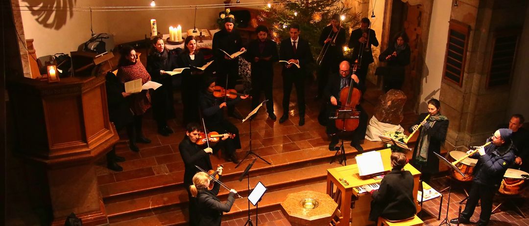 Ein Chor und ein Orchester führen ein Weihnachtskonzert in einer Kirche auf, mit Geigern, Cellisten und einem Pianisten. Der Chor liest Musik, und der Pianist sitzt. Ein Weihnachtsbaum und Kerzen schmücken den Altar.