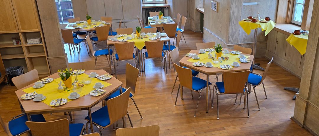 Ein Esszimmer mit mehreren Tischen und Stühlen, die jeweils mit Tellern, Tassen und gelben Tischdecken besetzt sind. Der Raum hat Holzböden und -wände, mit einem Fenster und einem Heizkörper sichtbar.