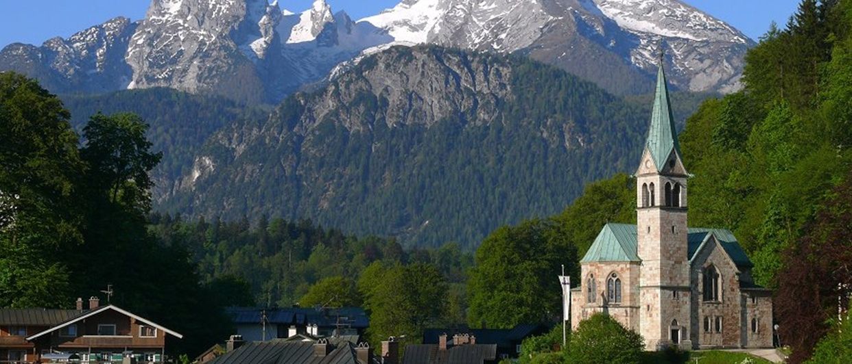 Eine kleine Stadtkirche mit einem grünen Turm steht zwischen grünen Bäumen und einem verschneiten Bergrücken.
