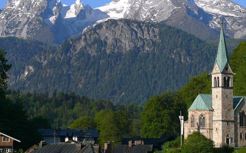 Eine kleine Stadtkirche mit einem grünen Turm steht zwischen grünen Bäumen und einem verschneiten Bergrücken.