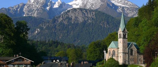 A small town church with a green steeple stands amidst green trees and a snowy mountain range.