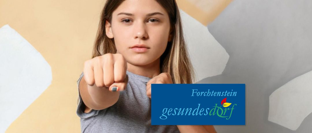 Ein Mädchen mit blonden Haaren, das ein graues T-Shirt trägt, macht eine Faust mit der rechten Hand und zeigt mit der linken Hand auf ihren Zeigefinger. Ein blaues Schild mit dem Text Forchtenstein und gesundesdorF befindet sich unten rechts.