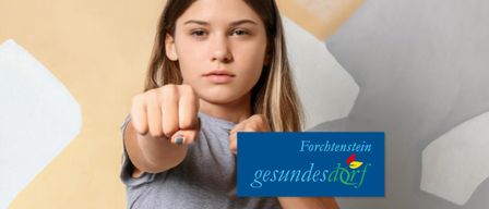 Ein Mädchen mit blonden Haaren, das ein graues T-Shirt trägt, macht eine Faust mit der rechten Hand und zeigt mit der linken Hand auf ihren Zeigefinger. Ein blaues Schild mit dem Text Forchtenstein und gesundesdorF befindet sich unten rechts.