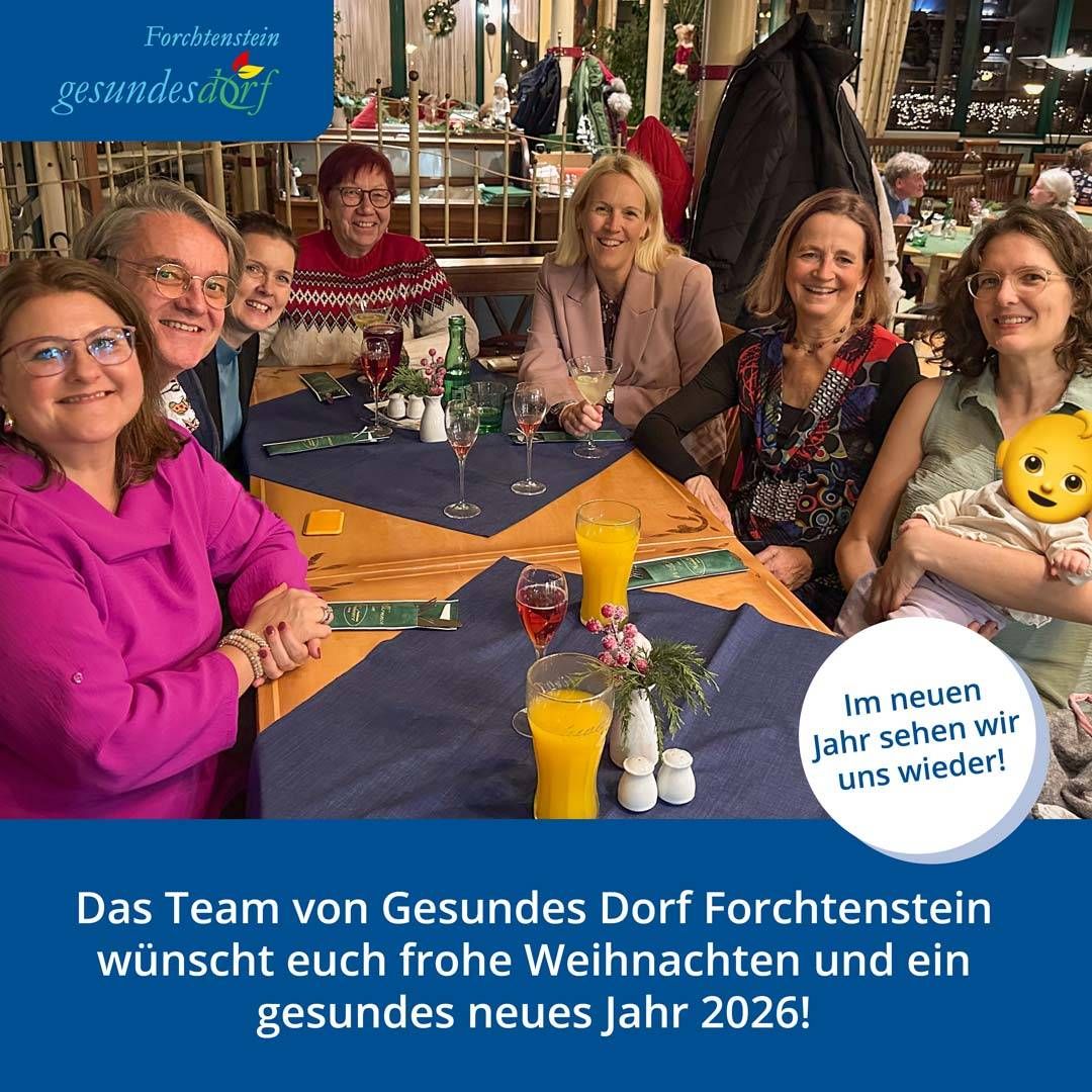 Eine Gruppe von Menschen sitzt an einem Tisch in einem Restaurant, lächelt und posiert für ein Foto. Auf dem Tisch stehen Getränke und eine Blumendekoration. Sie tragen Winterkleidung. Der Text am unteren Rand lautet: 'Das Team von Gesundes Dorf Forchtenstein wünscht euch frohe Weihnachten und ein gesundes neues Jahr 2026!' in Deutsch.