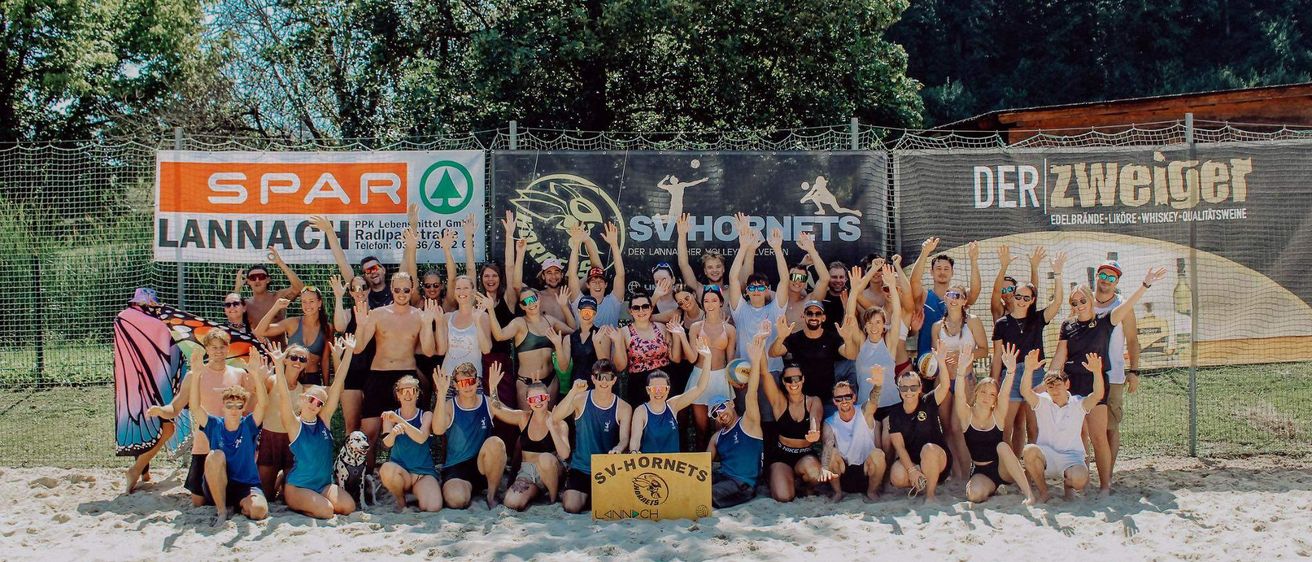 Eine Gruppe von Menschen posiert für ein Foto am Strand mit einem Banner, das SV Hornets steht. Sie alle lächeln und heben ihre Hände.