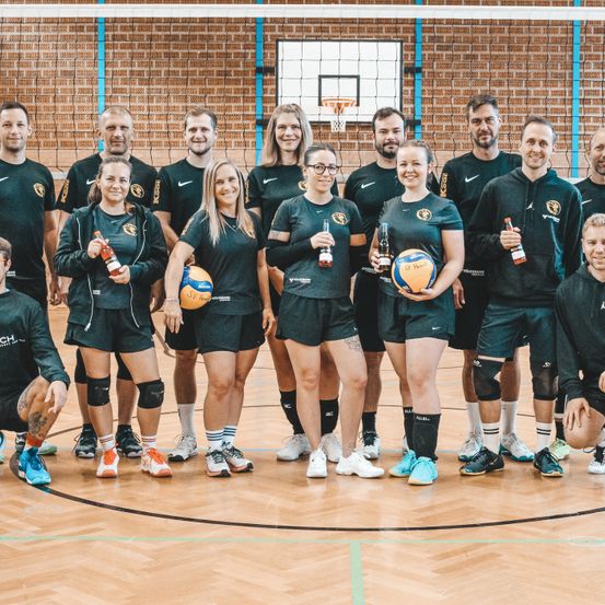 Eine Gruppe von Volleyballspielern in schwarzen Uniformen posiert mit Sportgetränken und Bällen für ein Foto in einer Turnhalle.