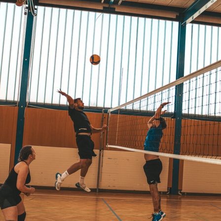 Drei Personen spielen Volleyball in einer Halle. Ein Mann springt und schlägt den Ball. Ein anderer Mann versucht, den Ball zu blocken. Eine Frau beobachtet.