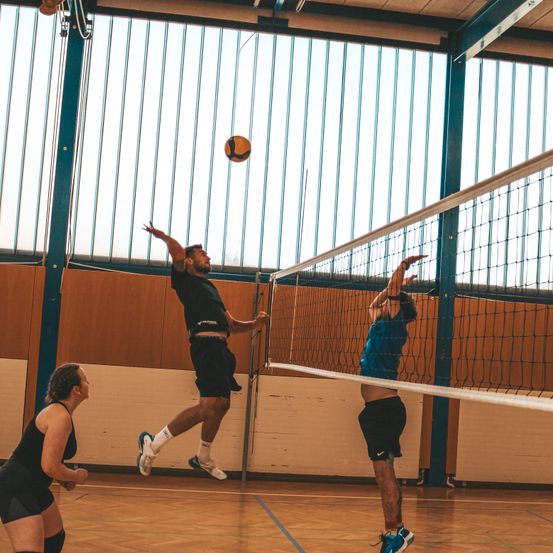 Drei Personen spielen Volleyball in einer Halle. Ein Mann springt und schlägt den Ball. Ein anderer Mann versucht, den Ball zu blocken. Eine Frau beobachtet.