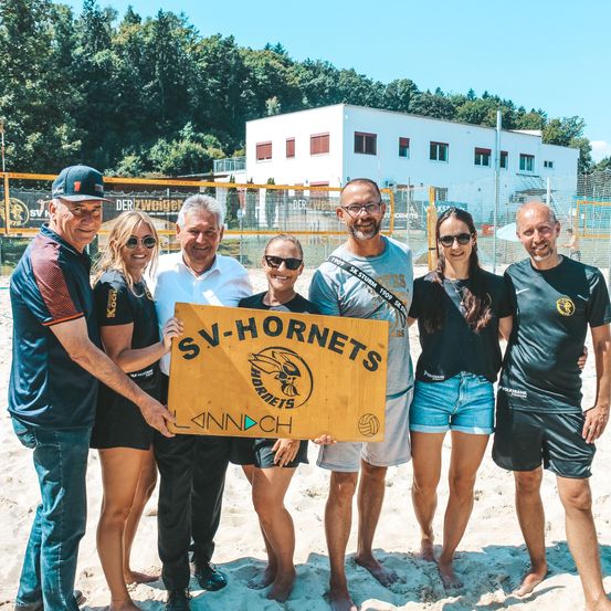 Eine Gruppe von Menschen steht am Strand mit einem Schild für SV-Hornets. Sie lächeln und posieren für ein Foto. Im Hintergrund steht ein weißes Gebäude und es gibt Bäume.