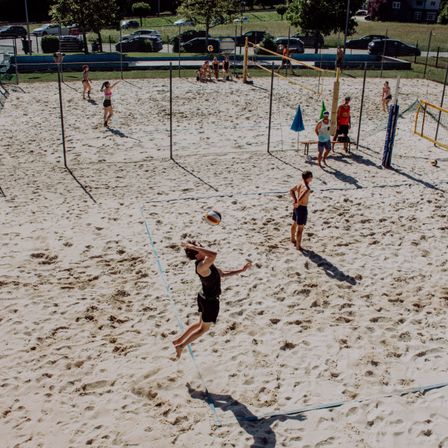 Mehrere Personen spielen Beachvolleyball auf einem Sandplatz. Einige springen, um den Ball zu schlagen. Das Spielfeld ist durch ein Netz geteilt.