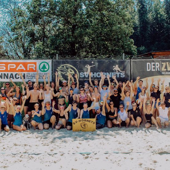 Eine Gruppe von Menschen in blauen und weißen Uniformen posiert für ein Foto am Strand, mit Bäumen und Bannern im Hintergrund.