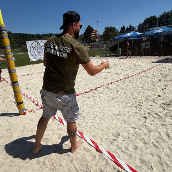 Ein Mann in einem grünen Shirt und Shorts spielt Beachvolleyball und steht auf dem Sandplatz. Ein blaues Zelt ist in der Nähe.