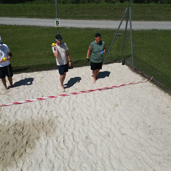 Drei Männer spielen ein Spiel auf einem sandigen Feld, zwei mit gelben Bällen, einer mit einem blauen Ball. Sie stehen hinter einem Netz.