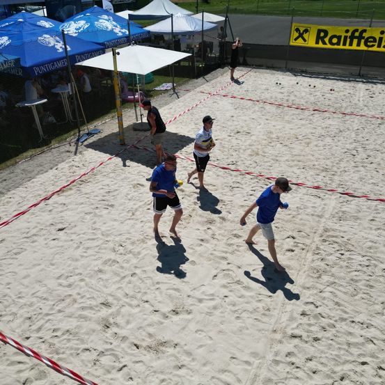Eine Gruppe von Menschen spielt Beachvolleyball auf einem sandigen Platz, wobei ein Netz das Feld teilt. Zuschauer beobachten das Spiel von Schirmen aus.