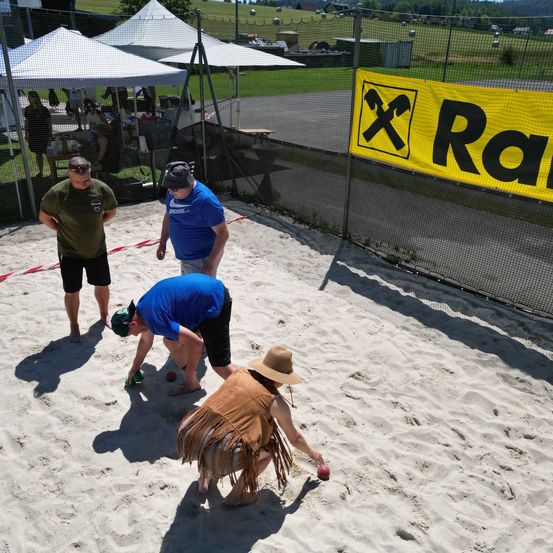 Eine Gruppe von Menschen spielt Strandspiele auf einem sandigen Feld mit Zelten und einem Zaun. Ein gelbes Banner mit dem Wort 'Ra' ist sichtbar.