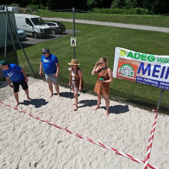 Vier Personen stehen in einem Sandbereich mit einem Schild, das Bau Ring zeigt. Einer hält einen Frisbee, während ein anderer auf ein Schild schaut. Dahinter sind ein Zaun, Autos und ein Rasenfeld sichtbar.