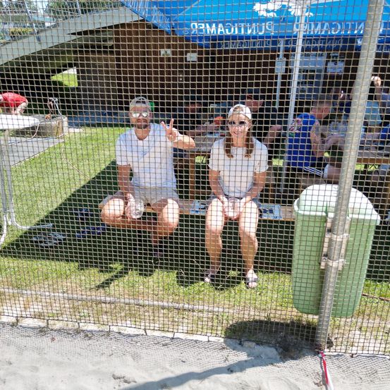 Ein Paar sitzt auf einer Bank hinter einem Zaun, mit einem blauen Zelt im Hintergrund. Der Mann macht ein Friedenszeichen und die Frau trägt Sonnenbrille. Andere Leute sind in der Nähe.