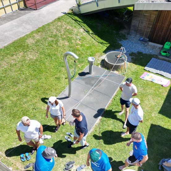 Eine Gruppe von Menschen versammelt sich auf einer Rasenfläche mit einer Wasserleitung und einer Dusche. Sie tragen Mützen und Turnschuhe, und einige sind barfuß. Handtücher und Schuhe sind auf dem Boden verstreut.
