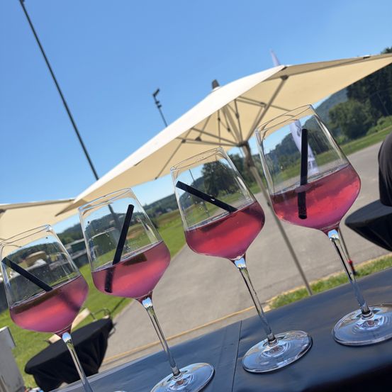 Vier Weingläser mit rosa Flüssigkeit und schwarzen Strohhalmen stehen auf einem Tisch unter weißen Sonnenschirmen. Im Hintergrund ist ein Rasenbereich und eine Straßenlaterne zu sehen.