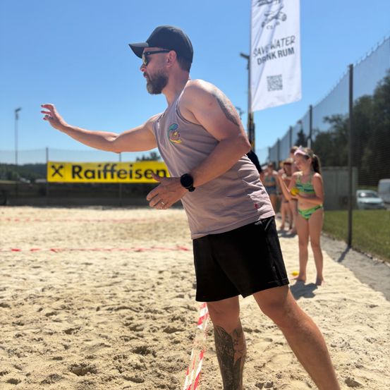 Ein Mann am Strand, der eine Kappe und Sonnenbrille trägt, bereitet sich darauf vor, einen Ball bei einem Beachvolleyballspiel zu werfen.