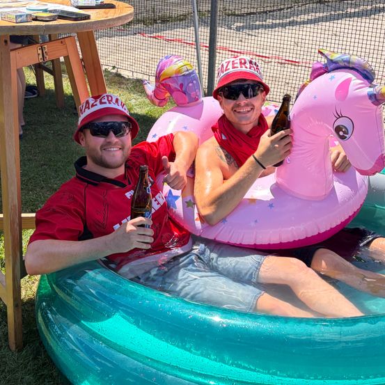 Zwei Männer sitzen in aufblasbaren Pool-Schwimmringen, einer hält ein Bier, trägt Hüte und Sonnenbrillen, und ein Mann in einem roten Shirt gibt ein Daumen hoch.