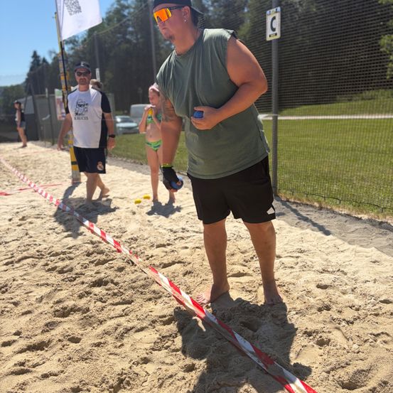 Ein Mann steht auf Sand, hält einen blauen Disc in der Hand, trägt eine Sonnenbrille und Shorts, mit anderen um ihn herum, einige gehen und andere stehen still.