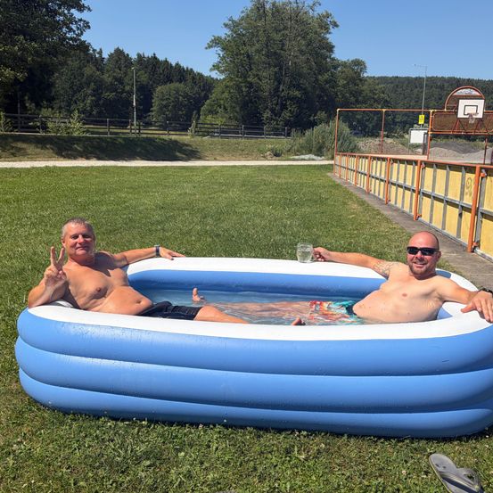 Zwei Männer entspannen sich in einem aufblasbaren Pool auf einem Grasfeld, mit einem Basketballkorb und Bäumen im Hintergrund.