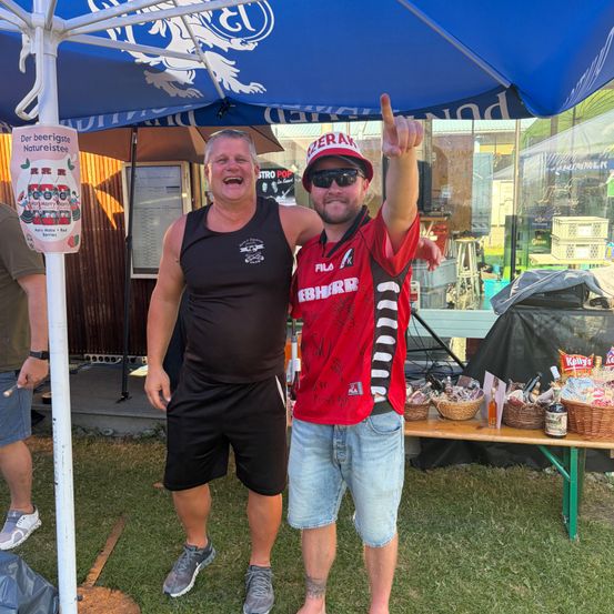 Zwei Männer posieren für ein Foto unter einem Zelt, einer trägt ein schwarzes Tanktop und der andere ein rotes Hemd. Sie lächeln und haben einen Tisch mit Gegenständen hinter sich.