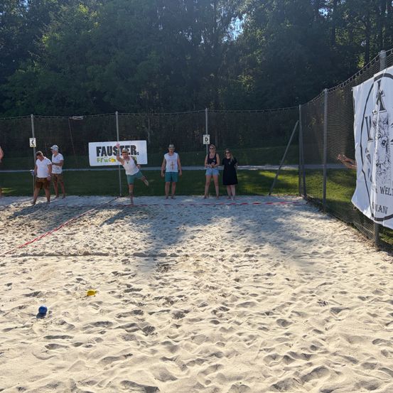 Eine Gruppe von Menschen spielt Beachvolleyball auf einem sandigen Gelände mit einem Zaun und Bannern im Hintergrund.