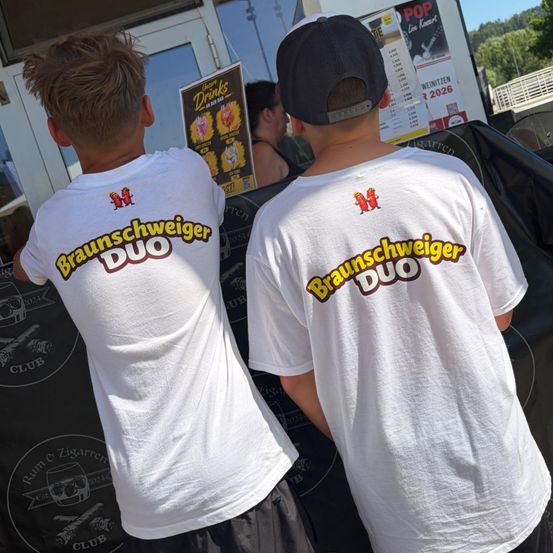 Zwei Jungen tragen identische weiße T-Shirts mit 'Braunschweiger Duo' auf der Rückseite. Sie tragen Mützen. Dahinter ist eine Speisekarte ausgestellt.