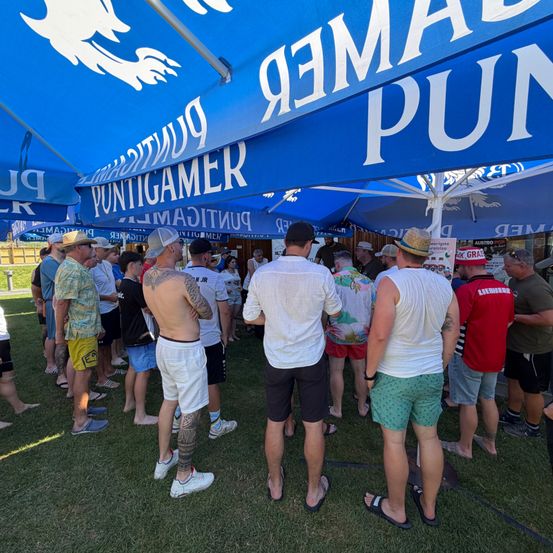 Eine Gruppe von Menschen in Freizeitkleidung wartet in einer Schlange unter einem blauen Zelt mit dem Wort 'PUNTIGAMER'. Sie stehen auf Gras, einige barfuß.