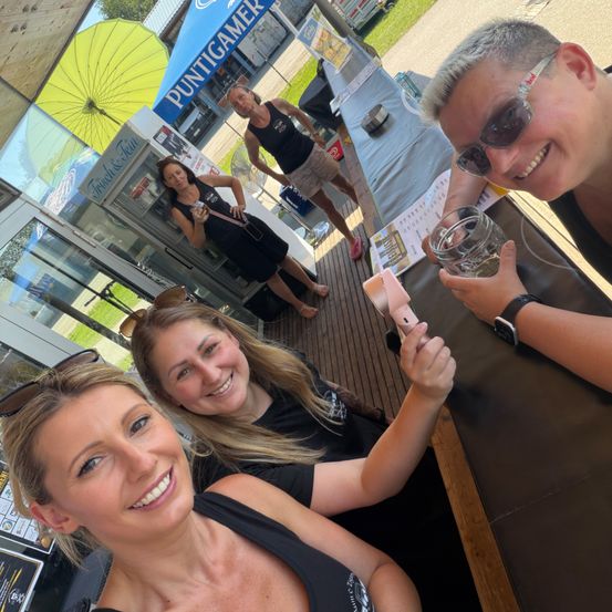 Vier Frauen lächeln und machen Selfies. Zwei sitzen an einem Tisch, eine hält ein Glas. Eine weitere Frau steht hinter ihnen.