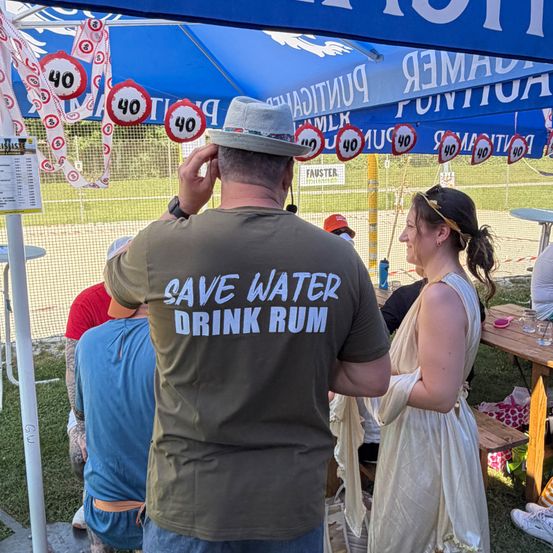 Ein Mann mit einem grünen T-Shirt, auf dem 'SAVE WATER DRINK RUM' steht, steht unter einem blauen Zelt. In der Nähe beobachtet eine Frau in einem weißen Kleid. Im Hintergrund eine Hecke, Tische und Schilder mit Zahlen und Buchstaben.