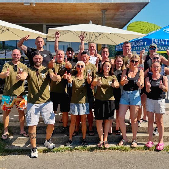 Eine Gruppe von Menschen in grünen Shirts und Shorts steht zusammen auf den Stufen eines Gebäudes. Einige tragen Sonnenbrillen und machen Gesten mit den Händen. Hinter ihnen sind Schirme und ein blaues Zelt.