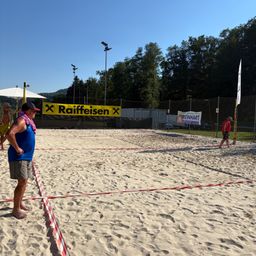 Ein Freiluft-Volleyballplatz mit einem gelben Banner, das Raiffeisen liest. Zwei Leute stehen, einer in einem roten Shirt und einer auf der gegenüberliegenden Seite.