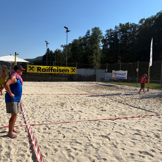 Ein Freiluft-Volleyballplatz mit einem gelben Banner, das Raiffeisen liest. Zwei Leute stehen, einer in einem roten Shirt und einer auf der gegenüberliegenden Seite.