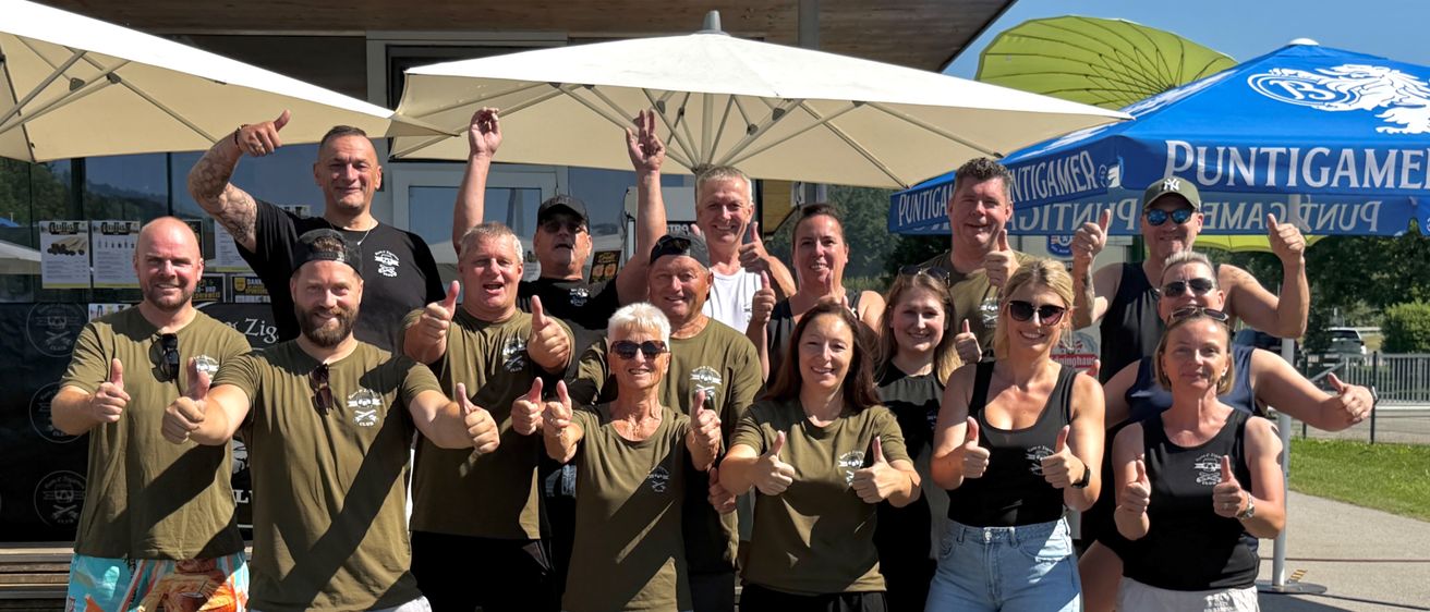 Eine Gruppe von Menschen in passenden grünen T-Shirts posiert für ein Foto, lächelt und gibt Daumen hoch. Sie stehen auf Stufen, mit Schirmen und einem Gebäude im Hintergrund.
