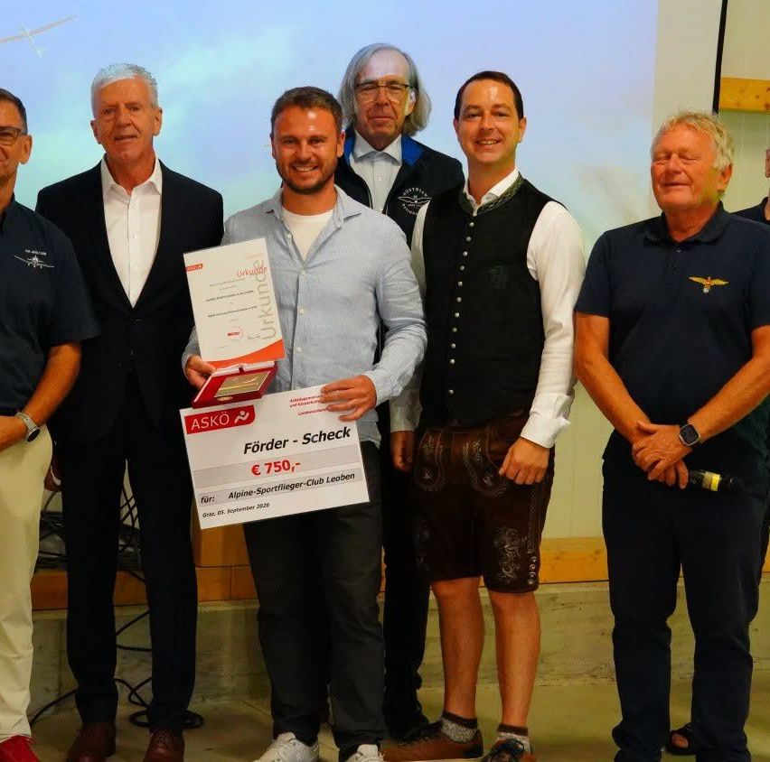 Eine Gruppe von Männern steht zusammen, einer hält ein Zertifikat und einen Scheck. Sie sind in verschiedenen Outfits gekleidet, darunter Anzüge, Westen und Freizeitkleidung. Der Mann in der Mitte hält ein Zertifikat und einen Scheck, während andere hinter ihm stehen.